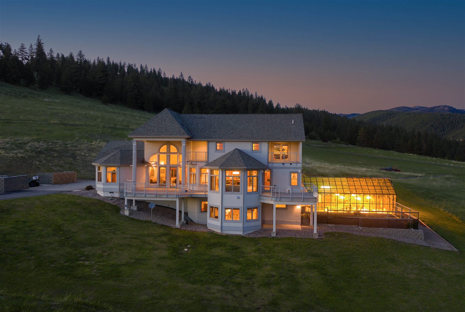 Twilight aerial of a large Montana home with glowing windows and greenhouse
